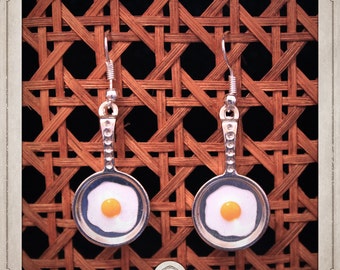 May include: A pair of silver earrings shaped like frying pans with a fried egg inside. The eggs are white with a yellow yolk.
