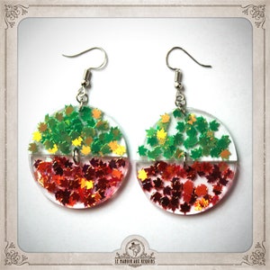 Puede incluir: Un par de pendientes con una base de resina transparente llena de hojas de purpurina verde y roja. Las hojas están dispuestas en forma de semicírculo, con las hojas verdes en la parte superior y las hojas rojas en la parte inferior.