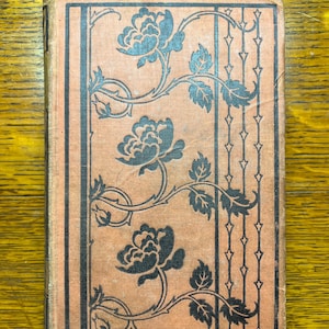 May include: A vintage book with a pink cover and black floral design. The design features roses and leaves with a repeating pattern of black diamonds along the spine.