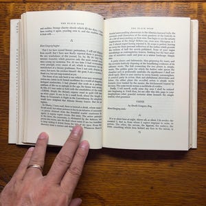 Puede incluir: Un libro abierto con texto, sostenido por una mano con un anillo de oro. El t&iacute;tulo del libro, "THE BLACK BOOK", es visible. Las p&aacute;ginas est&aacute;n amarillentas y el fondo es una superficie de madera.