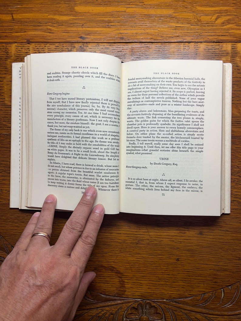 Puede incluir: Un libro abierto con texto, sostenido por una mano con un anillo de oro. El t&iacute;tulo del libro, "THE BLACK BOOK", es visible. Las p&aacute;ginas est&aacute;n amarillentas y el fondo es una superficie de madera.