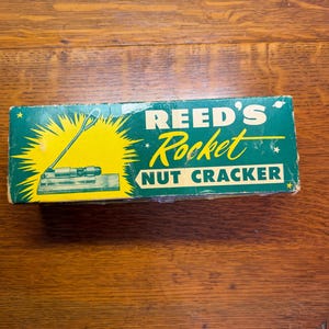 May include: A vintage box for Reed's Rocket Nut Cracker. The box is green with yellow accents and features an illustration of the nutcracker. The text on the box reads "REED'S Rocket NUT CRACKER".