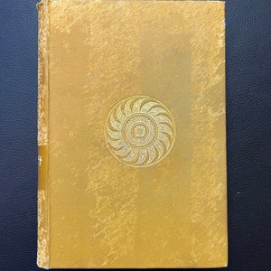 May include: A gold-colored book with a decorative design on the front cover. The design is a circular pattern with a central circle and radiating lines.