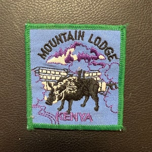 May include: A blue and green embroidered patch with the text "Mountain Lodge Kenya". The patch features a stylized image of a warthog in front of a building with a purple mountain in the background.