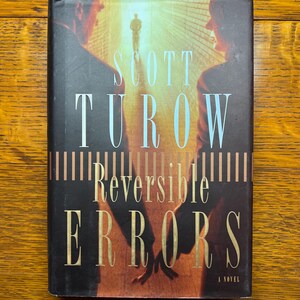 May include: A black hardcover book with a gold title, "Scott Turow Reversible Errors A Novel". The cover features a blurry image of two people walking towards a bright light.
