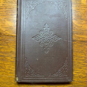 May include: A closed, antique book with a brown cover. The cover features an embossed, ornate design with corner and central embellishments. The spine has gold-colored accents. The book is rectangular and appears to be in good condition.