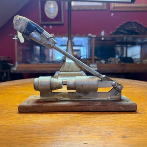 May include: An antique metal and wood device with a lever arm, likely a tool or instrument. The device is mounted on a wooden base and has a cylindrical component. The lever arm is wrapped with tape. The background is a room with antique decor.