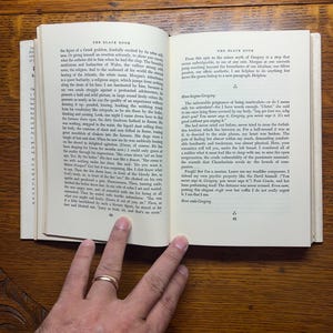 Puede incluir: Un libro abierto con texto en dos p&aacute;ginas enfrentadas. La p&aacute;gina izquierda tiene texto en una sola columna, mientras que la p&aacute;gina derecha tiene texto en dos columnas. El libro est&aacute; abierto sobre una superficie de madera, con una mano y un anillo de oro visibles.