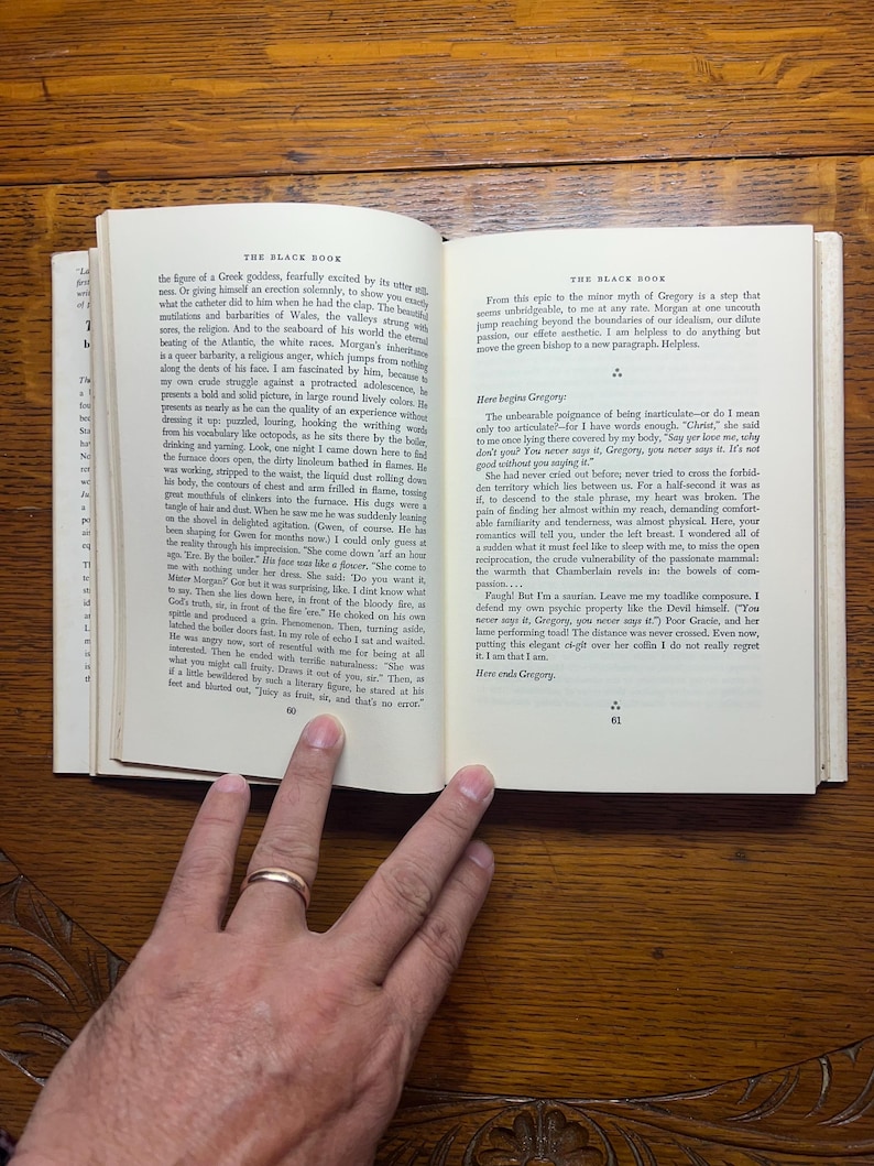 Puede incluir: Un libro abierto con texto en dos p&aacute;ginas enfrentadas. La p&aacute;gina izquierda tiene texto en una sola columna, mientras que la p&aacute;gina derecha tiene texto en dos columnas. El libro est&aacute; abierto sobre una superficie de madera, con una mano y un anillo de oro visibles.