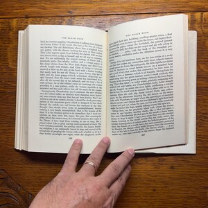 Puede incluir: Un libro abierto, titulado "THE BLACK BOOK", descansa sobre una superficie de madera. Las p&aacute;ginas est&aacute;n llenas de texto impreso. Una mano con un anillo de oro descansa sobre las p&aacute;ginas, oscureciendo parcialmente el texto. El libro est&aacute; abierto en las p&aacute;ginas 226 y 227.