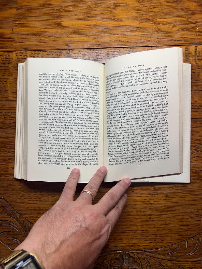 Puede incluir: Un libro abierto, titulado "THE BLACK BOOK", descansa sobre una superficie de madera. Las p&aacute;ginas est&aacute;n llenas de texto impreso. Una mano con un anillo de oro descansa sobre las p&aacute;ginas, oscureciendo parcialmente el texto. El libro est&aacute; abierto en las p&aacute;ginas 226 y 227.