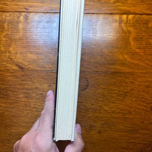 Puede incluir: Un libro con lomo negro y p&aacute;ginas blanquecinas es sostenido por una mano. Las p&aacute;ginas del libro est&aacute;n ligeramente amarillentas, lo que sugiere antig&uuml;edad. El libro descansa sobre una superficie de madera de tono marr&oacute;n c&aacute;lido.