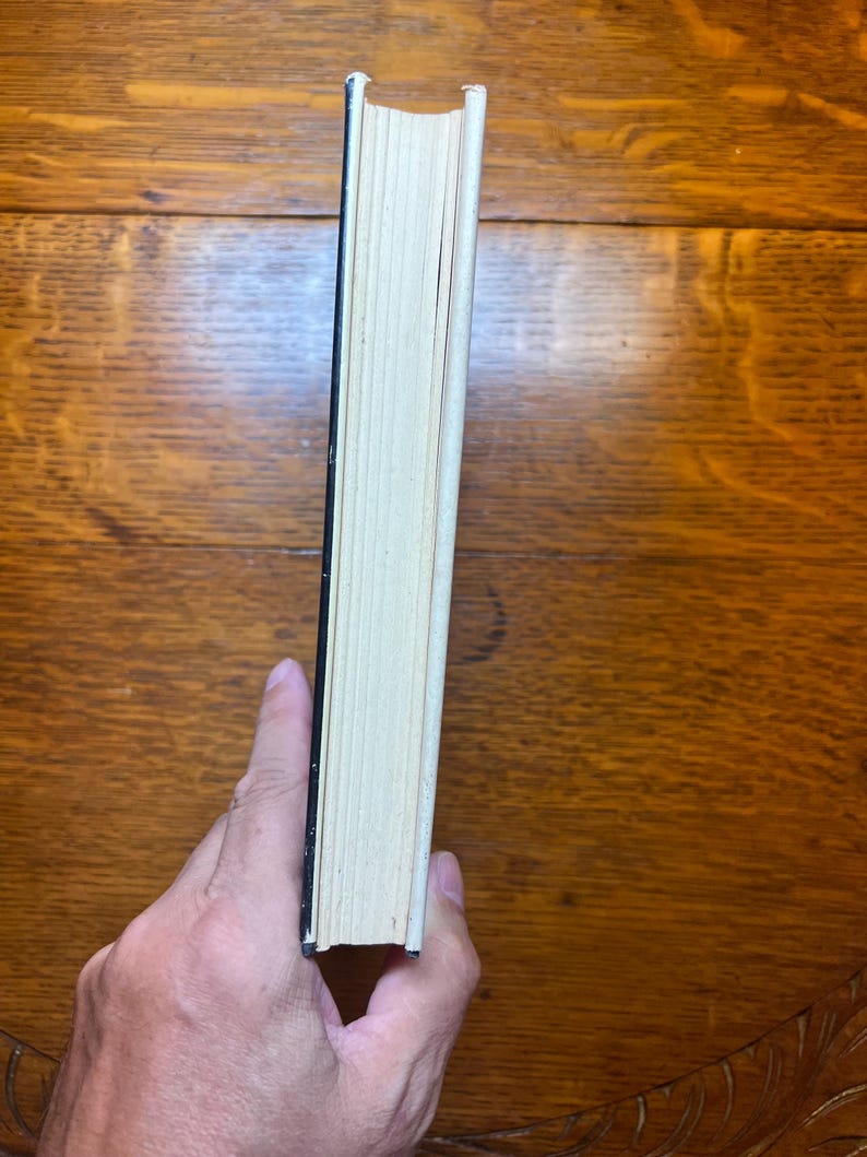 Puede incluir: Un libro con lomo negro y p&aacute;ginas blanquecinas es sostenido por una mano. Las p&aacute;ginas del libro est&aacute;n ligeramente amarillentas, lo que sugiere antig&uuml;edad. El libro descansa sobre una superficie de madera de tono marr&oacute;n c&aacute;lido.