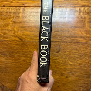 Puede incluir: Lomo de libro negro con el t&iacute;tulo "THE BLACK BOOK" en letras blancas. El nombre del autor, Durrell, est&aacute; en la parte superior. La editorial, Dutton, est&aacute; en la parte inferior. El libro est&aacute; sostenido por una mano.