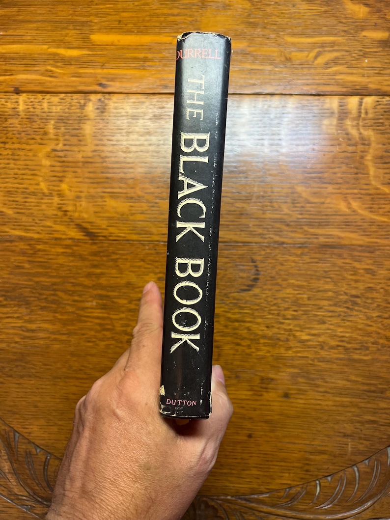 Puede incluir: Lomo de libro negro con el t&iacute;tulo "THE BLACK BOOK" en letras blancas. El nombre del autor, Durrell, est&aacute; en la parte superior. La editorial, Dutton, est&aacute; en la parte inferior. El libro est&aacute; sostenido por una mano.