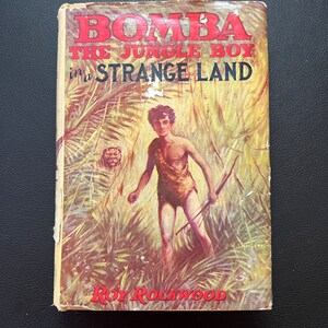 May include: A vintage book cover with the title "Bomba the Jungle Boy in a Strange Land" in red lettering. The cover features an illustration of a young boy in a jungle setting, holding a spear and walking through tall grass.