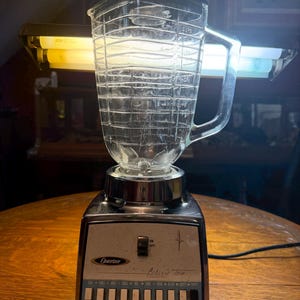 May include: A vintage Osterizer Galaxie Ten blender with a clear glass pitcher featuring embossed cup measurements. The base is black and white with a central on-off toggle switch, ten rectangular push-button speed controls, and a star graphic.