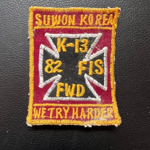 May include: A rectangular patch with a maroon background and gold trim. The patch features white text that reads "SUWON KOREA", "K-13", "82 FIS", and "FWD". The phrase "WE TRY HARDER" is also embroidered on the patch.