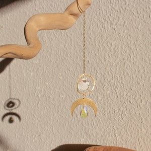 Könnte beinhalten: Ein goldfarbener Kristall-Sonnenfänger mit einem Halbmond und einem runden Kristall, der an einer Kette hängt. Der Sonnenfänger hängt an einem Stück Treibholz vor einer strukturierten beigefarbenen Wand.