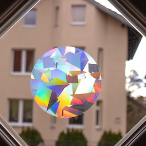 Op de afbeelding: Een ronde, iriserende sticker met een geometrisch patroon, aangebracht op een ruit. De sticker reflecteert licht in een regenboog van kleuren.