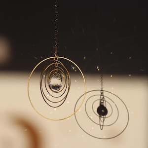 Pode incluir: Móvel dourado suspenso, composto por círculos concêntricos e um cristal central. A sombra do móvel é visível à direita. O fundo está desfocado, com uma paleta de cores neutras.