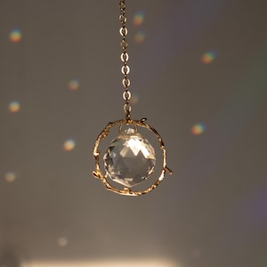 Könnte beinhalten: Eine Kristallkugel, die an einer goldenen Kette hängt, eingefasst in einen filigranen goldenen Drahtrahmen, der an Äste erinnert. Die Kristallkugel reflektiert das Licht und erzeugt regenbogenartige Muster auf dem Hintergrund.