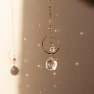 Peut inclure: Un attrape-soleil en métal doré avec un motif en spirale, comprenant deux sphères en cristal transparent et une boule de cristal facettée plus grande. L'attrape-soleil est suspendu à une chaîne délicate, créant un jeu de lumière et d'ombre.