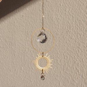 Könnte beinhalten: Ein Hängeornament mit Himmelsmotiv, das ein goldfarbenes Sonnenstrahlendesign und einen klaren Kristallkugel aufweist, die an einer Kette aufgehängt ist. Das Ornament enthält auch einen goldfarbenen Ring und einen kleineren Kristall. Der Hintergrund ist neutral.