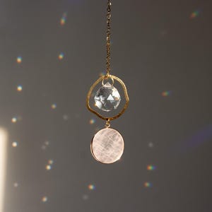Op de afbeelding: Een gouden kristallen zonenvanger met een heldere kristallen bol en een roze kristallen schijf die aan een ketting hangt.