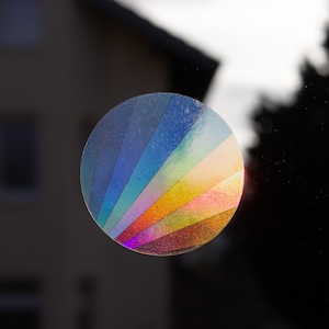 Könnte beinhalten: Ein runder, schimmernder Aufkleber mit einem Regenbogenmuster. Der Aufkleber besteht aus mehreren Schichten farbigem Films, die einen schimmernden Effekt erzeugen.
