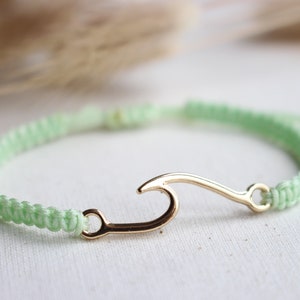 Könnte beinhalten: Ein hellgrünes geflochtenes Armband mit einem goldenen Wellen-Anhänger. Das Armband ist mit einem Schiebeknoten verstellbar.