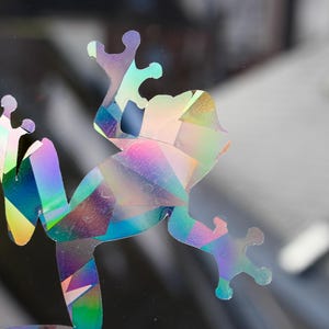 May include: A holographic frog sticker with a geometric design. The sticker has a rainbow color scheme, with shades of blue, green, pink, and yellow. The frog is in a side-facing pose, with its legs and arms extended.