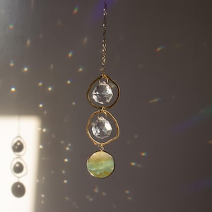 Puede incluir: Un atrapasol de cristal dorado con tres prismas de cristal transparente y un disco iridiscente verde y dorado que cuelga de una cadena dorada.