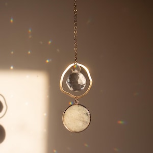 Könnte beinhalten: Eine goldene Kette mit einem klaren Kristall und einem weißen Steinanhänger. Der Anhänger ist rund und hat eine glatte Oberfläche.