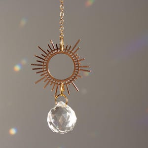 Op de afbeelding: Een gouden zonnestralenhanger met een helder kristal dat eraan hangt. De hanger hangt aan een gouden ketting.