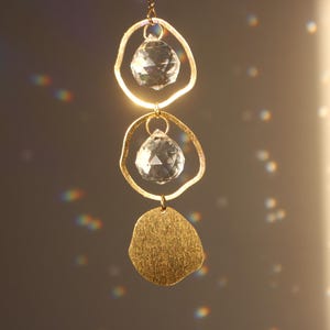 Puede incluir: Un adorno colgante dorado con dos colgantes de cristal transparente y un dije dorado texturizado de forma irregular. El adorno está suspendido de una delicada cadena, reflejando la luz y creando un efecto brillante. El fondo es de un tono suave y neutro.