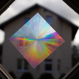 Op de afbeelding: Een regenboogkleurige, iriserende sticker in diamantvorm met een radiaal kleurenschema, aangebracht op een raam met een wazig zicht op een huis op de achtergrond.