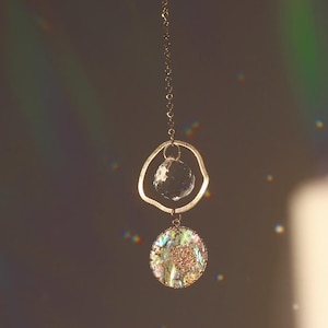 Könnte beinhalten: Ein hängender Sonnenfänger mit einem durchsichtigen Kristallkugel und einem schillernden Abalone-Muschel-Anhänger. Der Sonnenfänger wird von einer zarten goldfarbenen Kette gehalten, die einen schimmernden Effekt erzeugt. Der Hintergrund ist ein weicher, neutraler Ton mit Regenbogenlicht.