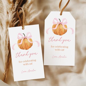 Puede incluir: Dos etiquetas de regalo blancas con un diseño de calabaza en acuarela y cinta rosa. Las etiquetas dicen "¡gracias por celebrar con nosotros!" y "Love, Amelia" en escritura rosa. Cada etiqueta tiene una cinta marrón.