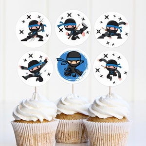 Puede incluir: Seis toppers redondos para cupcakes con un ninja de dibujos animados con un traje negro y una cinta azul. El ninja sostiene una espada y estrellas ninja. Los toppers están en forros de cupcakes blancos.