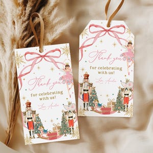 May include: Two white gift tags with a watercolor design. Each tag features a pink ribbon, text that reads "Thank you for celebrating with us!", and illustrations of a ballerina, nutcracker, and Christmas tree. Gold accents.