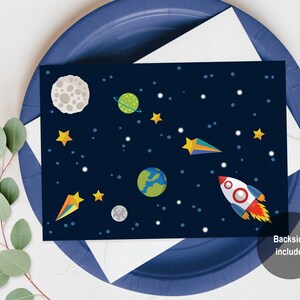 May include: A blue card with a space-themed design. The card features a rocket, planets, stars, and a crescent moon. The text "Backside Included" is printed on the bottom right corner of the card.