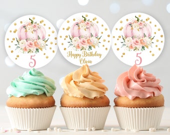 May include: Three cupcakes with pink, yellow, and green frosting. Each cupcake has a round white topper with a pink number 5 and a gold glitter pumpkin with pink flowers. The middle topper says "Happy Birthday Olivia".