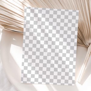 May include: A blank white card with a checkered pattern sits on a beige surface with a dried palm leaf behind it.