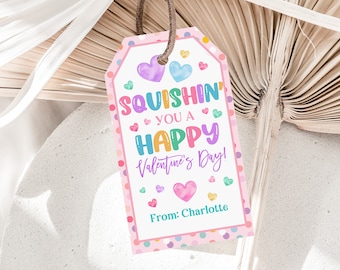 Pode incluir: Uma etiqueta de presente de Dia dos Namorados com uma borda rosa e bolinhas coloridas. A etiqueta diz "Squishin' You a Happy Valentine's Day!" com corações e o nome "Charlotte" na parte inferior. Uma corda marrom está presa no topo.
