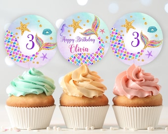 May include: Three mermaid-themed cupcake toppers with purple, blue, and gold glitter. The toppers feature a mermaid tail, a star, and the number 3. One topper also says "Happy Birthday Olivia".