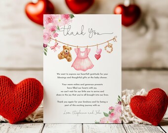 May include: Thank you card with a pink floral design and a line of baby items hanging on a clothesline. The items include a teddy bear, a pink dress, a heart, and a bracelet. The text on the card reads "Thank You".