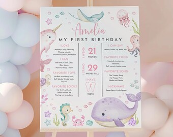 May include: A pink and blue illustrated first birthday chart for a baby girl named Amelia. The chart includes details about her weight, height, favourite foods, songs, and toys. The chart features a whale, a narwhal, a seahorse, and other sea creatures.