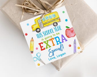 Puede incluir: Una etiqueta de regalo con una ilustración de acuarela de un autobús escolar amarillo, una manzana, un lápiz, unas tijeras y un crayón. El texto dice "THIS SCHOOL YEAR is going to be EXTRA Special! Love, Logan."