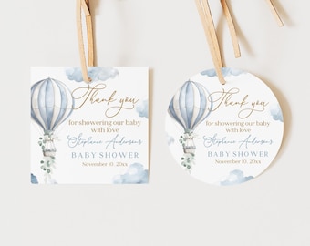 Può includere: Due etichette regalo per baby shower. Una è quadrata, l'altra rotonda. Entrambe presentano un disegno di mongolfiera blu e bianca con il testo "Thank you" e "Baby Shower". Le etichette sono legate con un nastro marrone chiaro.